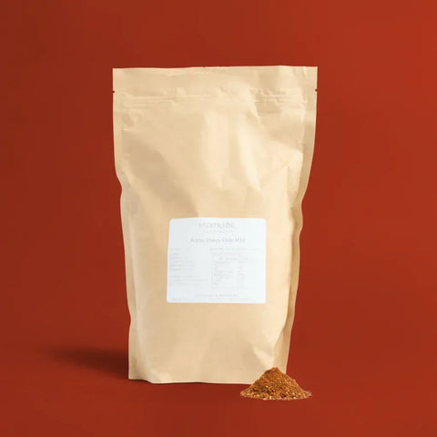 Beige paper bag of Ancho Flakes with a small pile of flakes in front on a red background