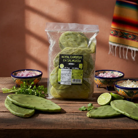 Packaged green nopal cactus on a wooden table with ingredients and a colorful towel in the background.