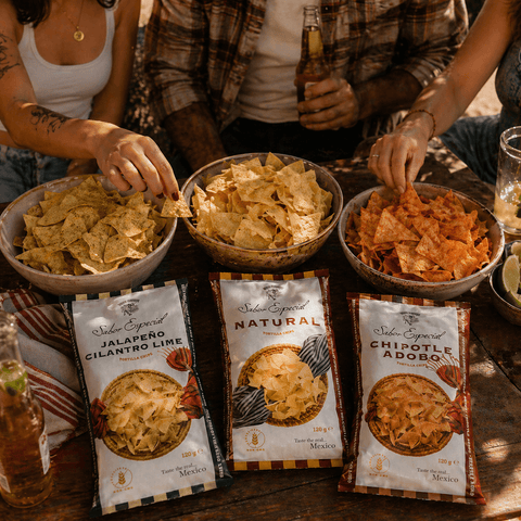 Three people are enjoying bowls of tortilla chips with Sabritas packaging on a wooden table.