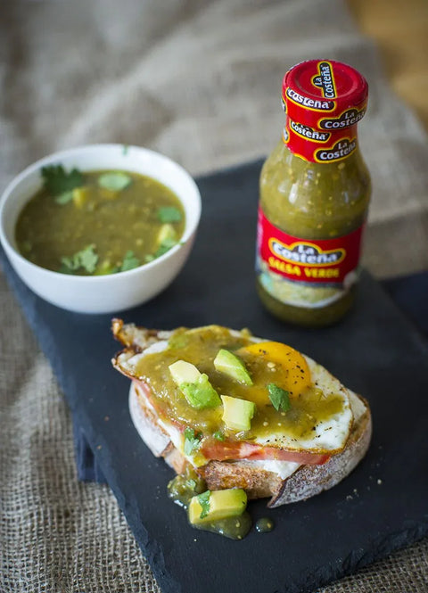 Bottle of La Costena salsa verde with a bowl of green salsa and a sandwich on a black slate board.