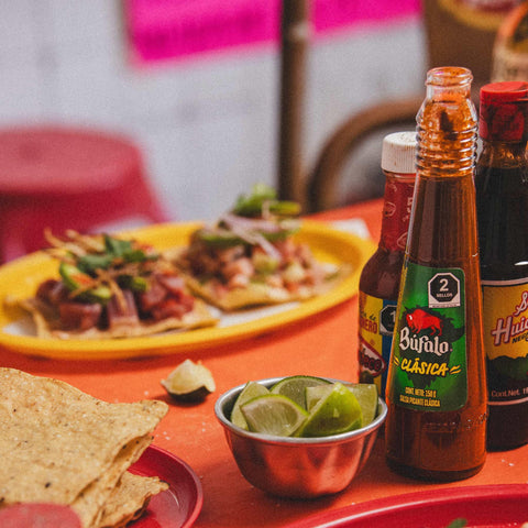 Bufalo Hot sauce bottle with a plate of nachos and a bowl of limes on a red table.