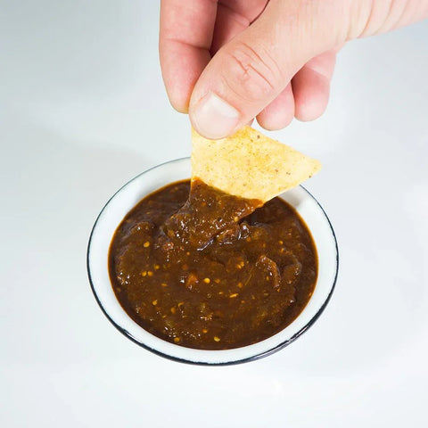 Hand dipping a tortilla chip into a bowl of ranchera salsa on a light background